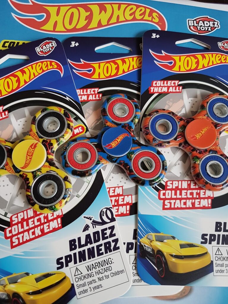 Three Hot Wheels-themed Bladez Spinnerz fidget spinners in colourful packaging, each with a round Hot Wheels logo in the centre. The packages are blue, white, and red, and feature a yellow racing car illustration and warnings for small parts.