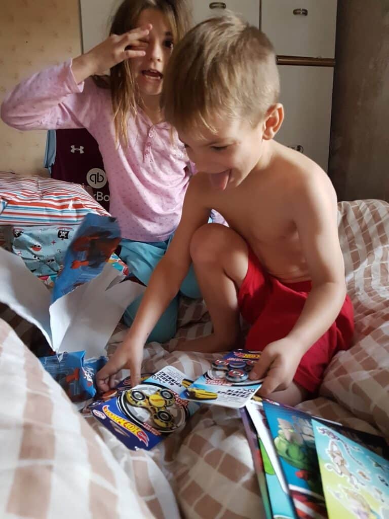 A smiling young boy in red shorts examines toy packages and books on a bed, with torn wrapping paper beside him. A girl in pink pyjamas is behind him, making a playful gesture with her fingers in front of her face. Both appear excited and happy.