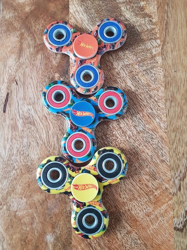 Four colourful fidget spinners with Hot Wheels logos are arranged vertically on a wooden surface. The spinners have blue, red, and yellow designs, each with three outer bearings and a central bearing.
