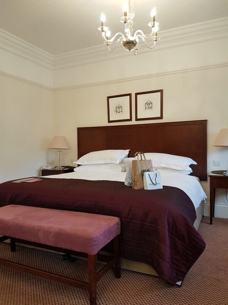 A neatly made hotel bed with white sheets, a burgundy throw, and two shopping bags on top. There are matching bedside tables with lamps, framed pictures above the headboard, a chandelier overhead, and a pink upholstered bench at the foot of the bed.