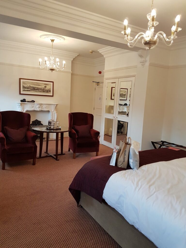 A cosy hotel room with cream walls and carpet, two red high-backed armchairs by a round table, a bed with white and burgundy bedding, chandeliers, a mantelpiece, and double doors with mirrors. Shopping bags are on the bed, and a suitcase rack is by the wall.