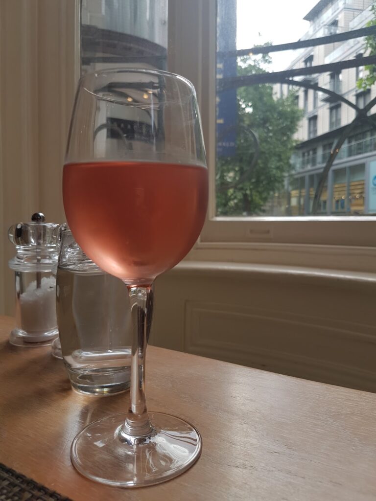 A glass of rosé wine sits on a wooden table beside a glass of water and salt and pepper shakers. Behind them, a window reveals a city street with trees, shopfronts, and a building under a cloudy sky.