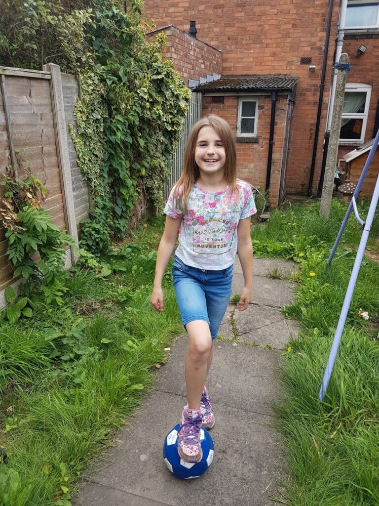 A smiling young girl with straight light brown hair stands on a garden path, wearing a floral t-shirt, blue shorts, and purple trainers. She balances one foot on a blue and white football. The garden has grass, overgrown plants, and a brick house in the background.