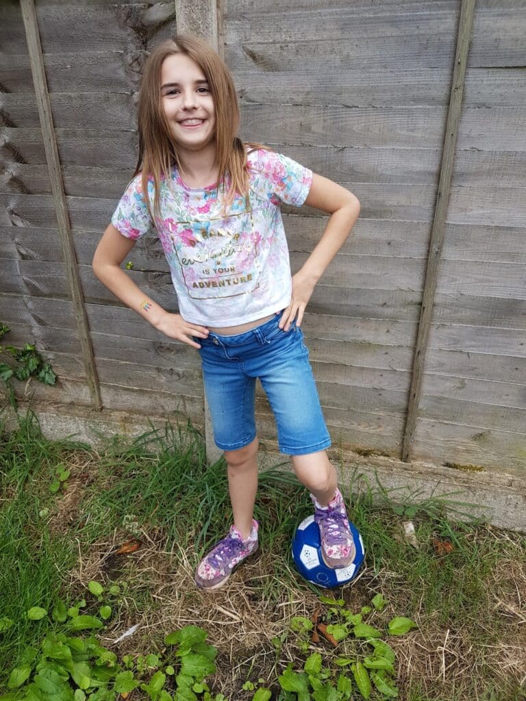 A smiling girl with long brown hair stands outdoors on grass, wearing a floral T-shirt, blue denim shorts, and floral trainers. She rests one foot on a blue and white football, with a wooden fence in the background.