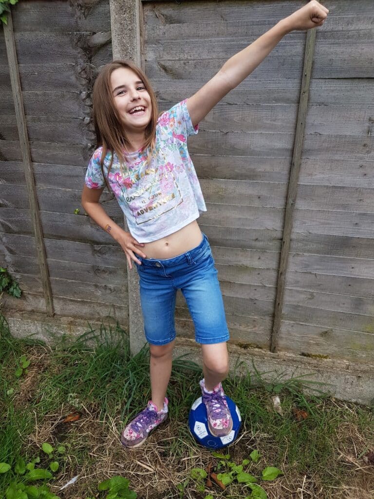 A young girl stands outside on grass near a wooden fence, smiling joyfully with one arm raised in the air. She wears a colourful t-shirt, blue denim shorts, and floral shoes. One foot rests on a blue-and-white football.