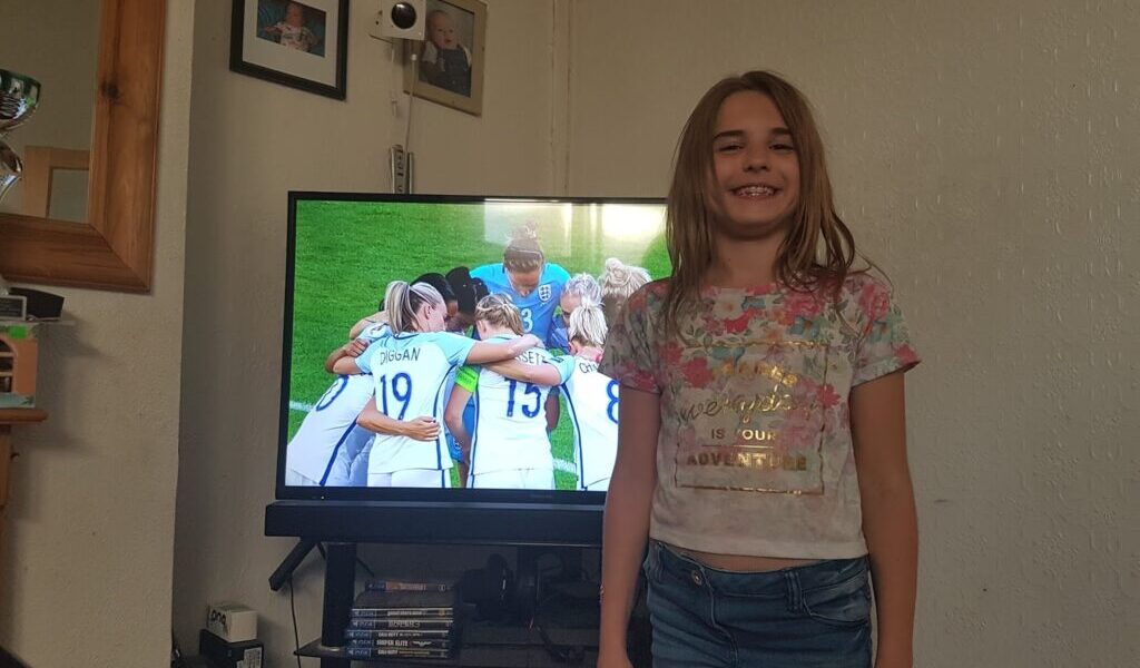 A smiling young girl with long hair, wearing a floral t-shirt and jeans, stands in a living room. Behind her, a TV shows a women’s football team huddling on a green pitch. The room has framed photos, a mirror, and various electronics visible.