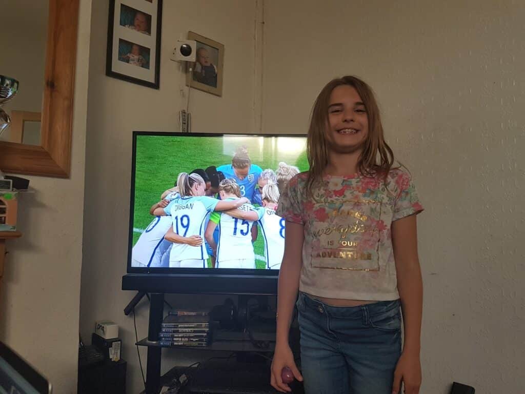 A smiling girl with long hair and a floral t-shirt stands indoors, in front of a TV showing a women’s football team huddled together on a pitch. The room has a mirror, framed photos, a white wall, and a shelf with DVDs below the TV.