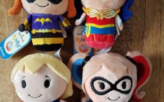 Four plush dolls of DC Comics female superheroes—Batgirl, Wonder Woman, Supergirl, and Harley Quinn—lie on a wooden surface. Each doll has a smiling face, costume details, and a tag reading "itty bittys." The background is brown wood.