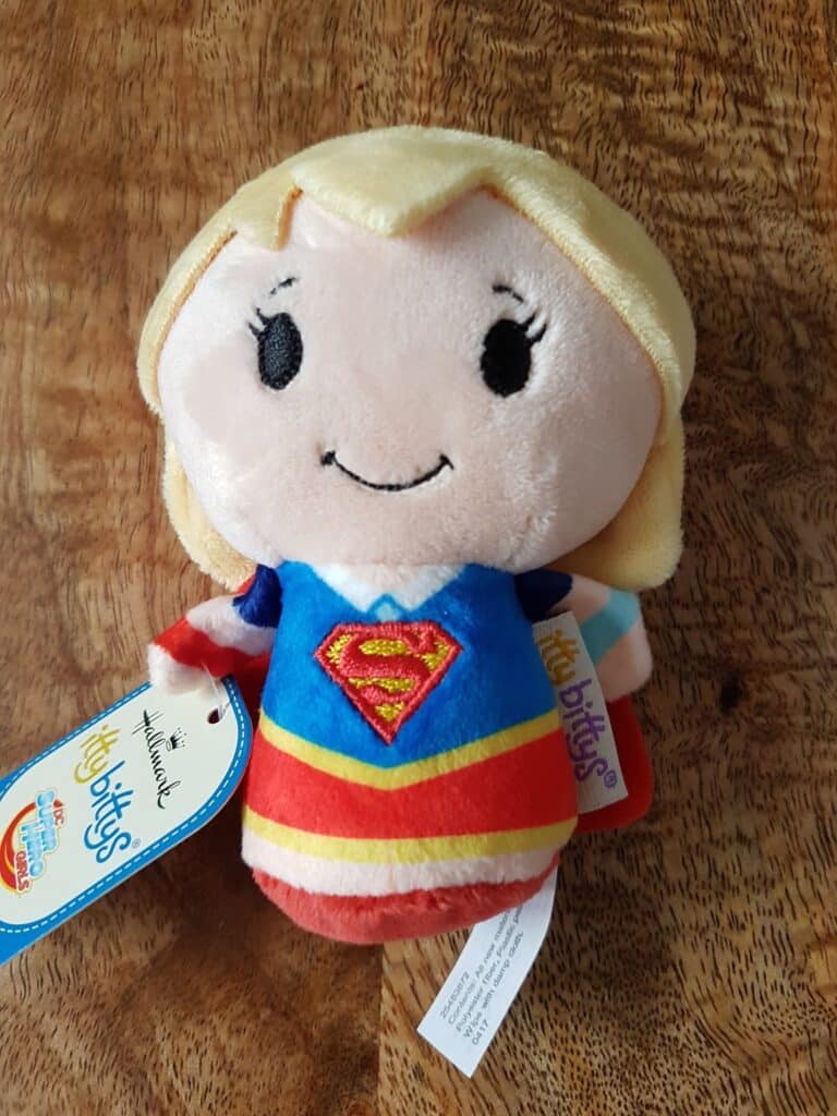 A plush toy of Supergirl with blonde hair, black embroidered eyes, and a smile. She wears a blue suit with a red and yellow “S” logo, a red skirt, and a yellow belt. The toy has tags attached and rests on a wooden surface.