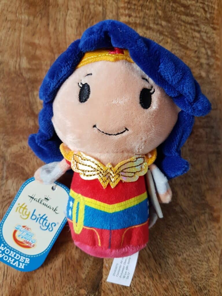 A plush Hallmark itty bittys doll styled as Wonder Woman stands on a wooden surface. The doll has blue hair, a red and gold headband, a gold emblem on her chest, a red top, blue skirt, and holds a tag that reads "Wonder Woman.