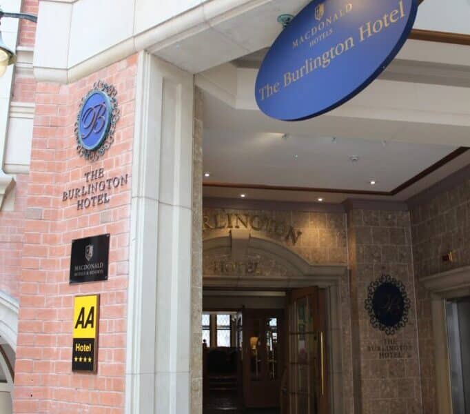 Entrance to The Burlington Hotel with a blue oval sign above the doorway and another plaque on the brick wall. The entrance is covered, leading to glass doors inside. An AA Hotel rating badge is also visible on the wall near the entrance.