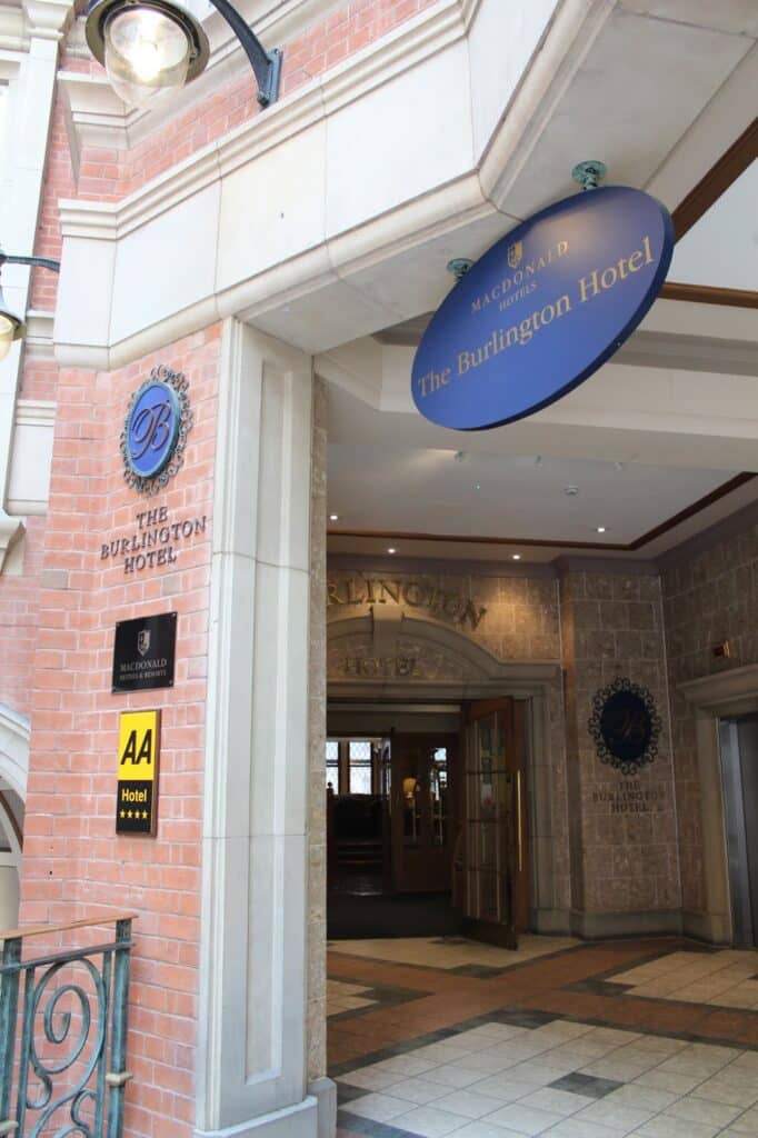 Entrance to The Burlington Hotel with a large blue oval sign overhead, a round wall plaque, and an AA Hotel sign beside the door. The hotel’s entrance is framed by brick and stone, and glass doors are visible inside.