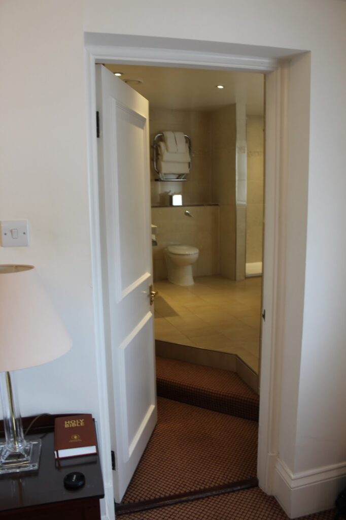A slightly open white door reveals a bathroom with a toilet, beige walls, and a towel rail. The foreground shows part of a bedroom with a lamp, a bedside table, and a book titled "Holy Bible" on the table. The floor transitions from carpet to tiles.