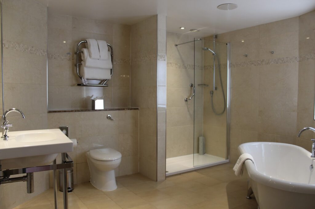 Modern bathroom with beige tiles, a glass-enclosed shower, a white freestanding bath, a white basin with chrome fittings, and a toilet. Towels are neatly stacked on a wall-mounted rack above the toilet. The space looks clean and well-lit.