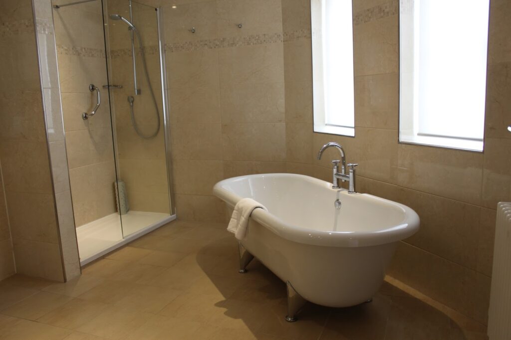 A modern bathroom with beige tiled walls and floors, featuring a glass-enclosed shower with chrome fittings, a white freestanding clawfoot bath with a towel draped over the edge, and two large frosted windows letting in natural light.