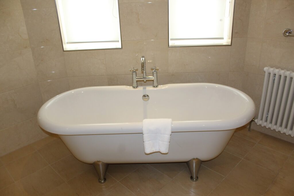 A white, freestanding bathtub with metal feet stands in a tiled bathroom corner. A white towel hangs over the bath’s edge. Two small windows with blinds are above the bath, and a white radiator is visible to the right. The room has beige tiled walls and floor.