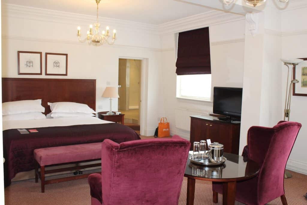A hotel room with a double bed, purple bench, two purple chairs around a glass table with a tray of glasses and water, TV on a dark wooden stand, window with dark blind, paintings on the wall, chandelier, and a door to an en suite bathroom.