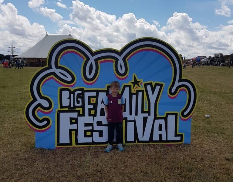 A young boy stands smiling in front of a large colourful sign that says “Big Family Festival” on a grassy field. There is a white marquee and people in the background under a partly cloudy blue sky.