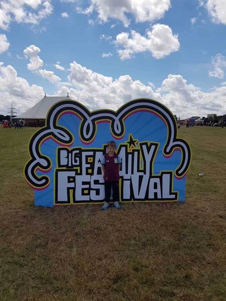 A young child stands smiling in front of a large, colourful "Big Family Festival" sign on a grassy field, with a white event marquee, people, and vehicles visible in the background under a blue sky with scattered clouds.