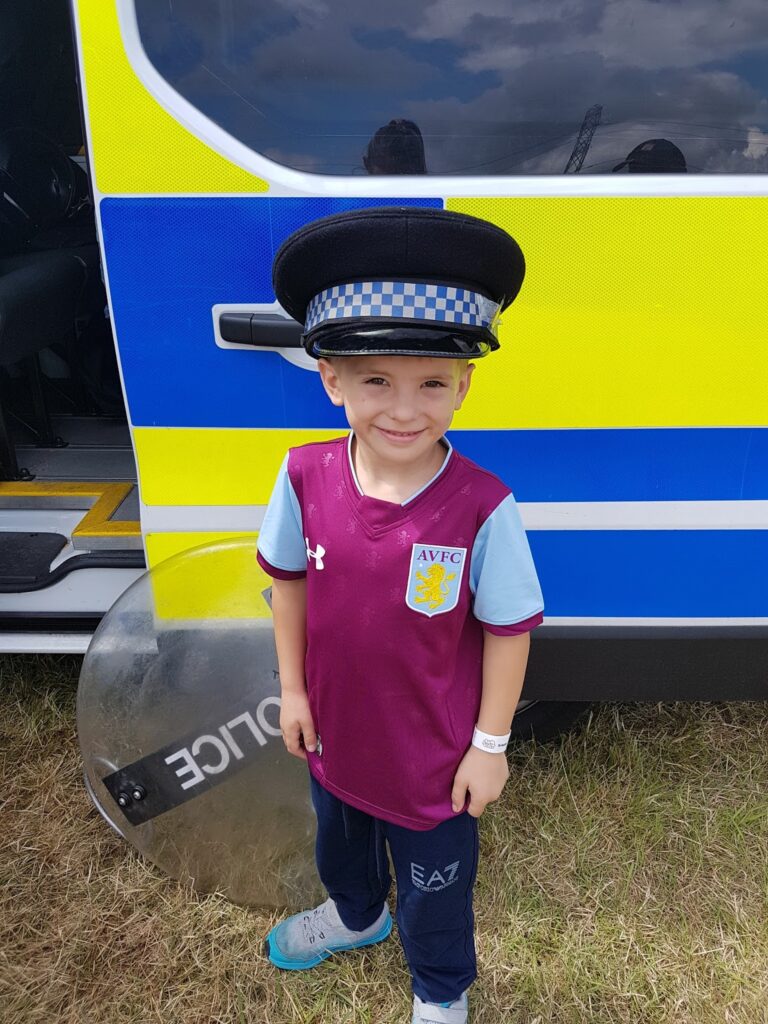 A young boy in an Aston Villa football shirt stands outside a police van, smiling. He is wearing a large police hat that covers his ears, and a police riot shield is beside him. The background shows the side of the police van with bright yellow and blue markings.