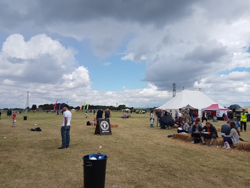A grassy field with groups of people sitting on hay bales and walking around at an outdoor event. A large white marquee and colourful flags are in the background. Rubbish bins are scattered around, and the sky is cloudy with patches of blue.