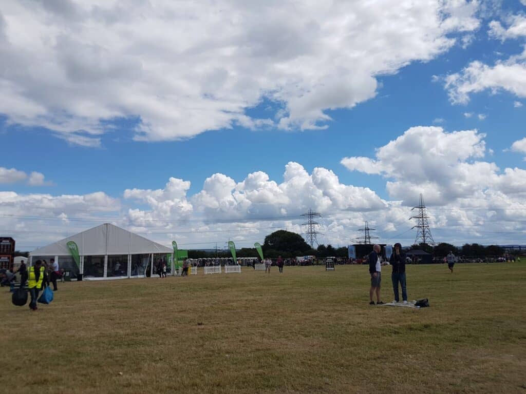 A large grassy field with scattered people, a white event marquee, and green flags. The sky is mostly cloudy with patches of blue. Power lines and pylons are visible in the background, and groups of people are walking or standing around the open space.