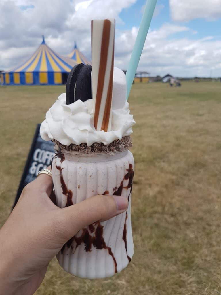 A hand holds a glass milkshake topped with whipped cream, a chocolate biscuit, marshmallow, wafer sticks, and chocolate syrup. The background shows a grassy field with a blue-and-yellow circus tent and cloudy sky.