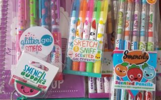 A collection of colourful scented stationery items, including gel pens, crayons, pencils, and sketch pads, all featuring cute cartoon fruit faces. The items are arranged neatly on a wooden surface with a bright green sensory pop-it mat at the bottom.