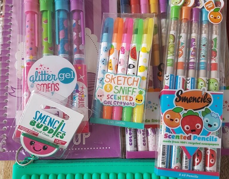 A collection of colourful scented stationery items, including gel pens, crayons, pencils, and sketch pads, all featuring cute cartoon fruit faces. The items are arranged neatly on a wooden surface with a bright green sensory pop-it mat at the bottom.