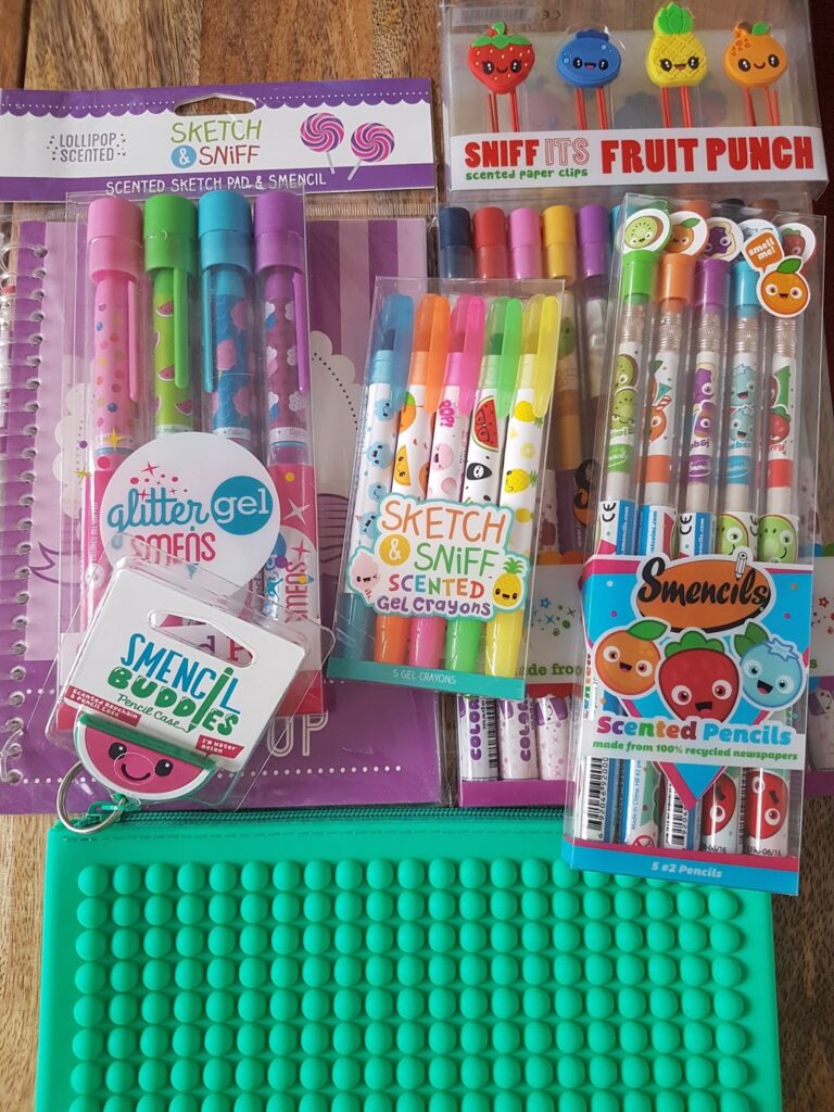 A collection of colourful scented stationery items, including gel pens, gel crayons, pencils, and markers, all with playful packaging featuring fruit and cartoon faces. The items are arranged on a green pop-it fidget toy and a purple notebook with spiral binding.