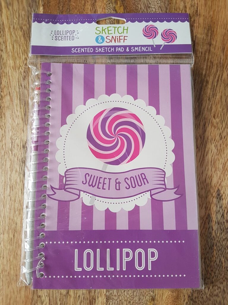 A purple spiral-bound notebook with a "Sweet & Sour" lollipop design on the cover. The packaging reads "Sketch & Sniff Scented Sketch Pad & Stencil" and "Lollipop Scented." The notebook rests on a wooden surface.