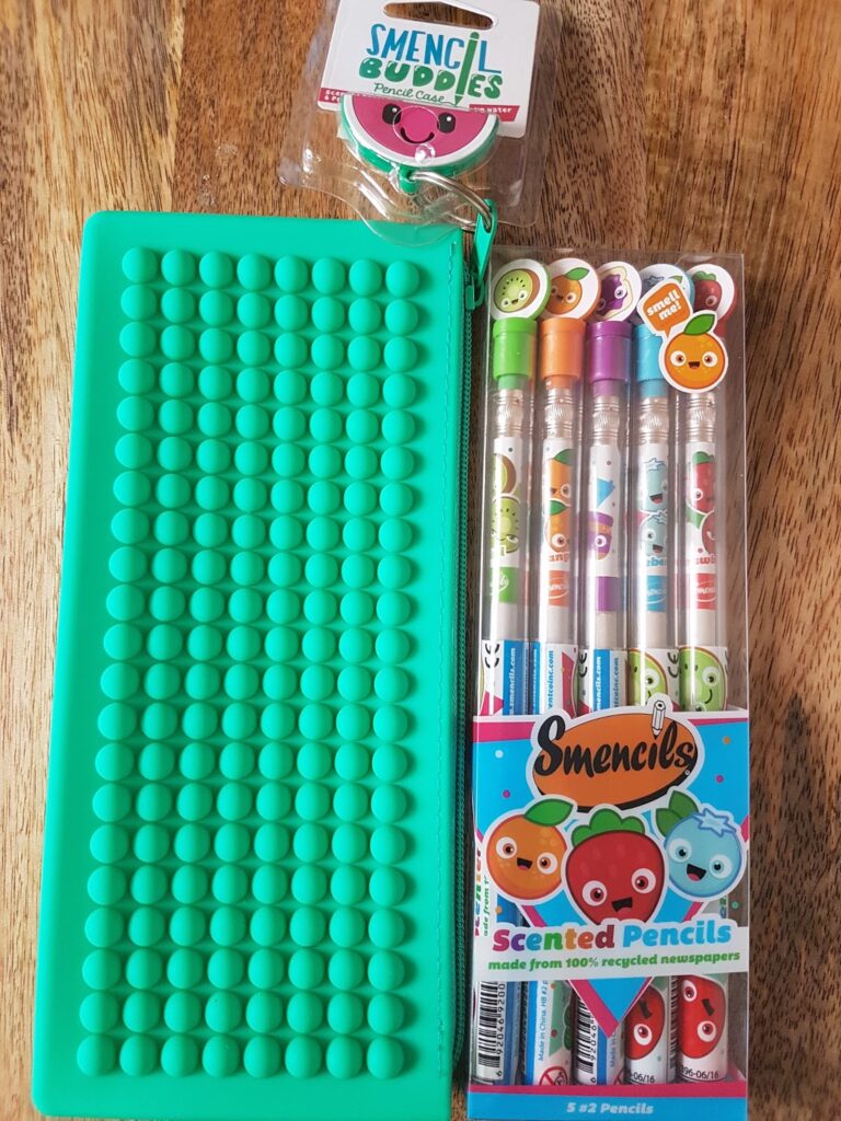 A teal silicone pop-it toy lies on a wooden surface next to five scented pencils in clear packaging featuring colourful cartoon fruits and labelled “Smencils.” Above is a small container labelled “Smencil Buddies” with a smiling watermelon character on the lid.