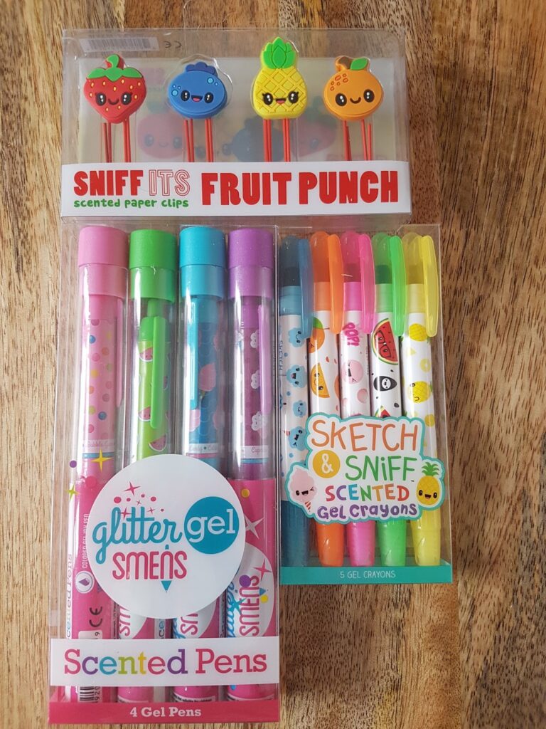 A set of colourful stationery: four fruit-themed scented paper clips, four glitter gel scented pens with cute designs, and five scented gel crayons with fruit faces, all in clear plastic packaging on a wooden surface.