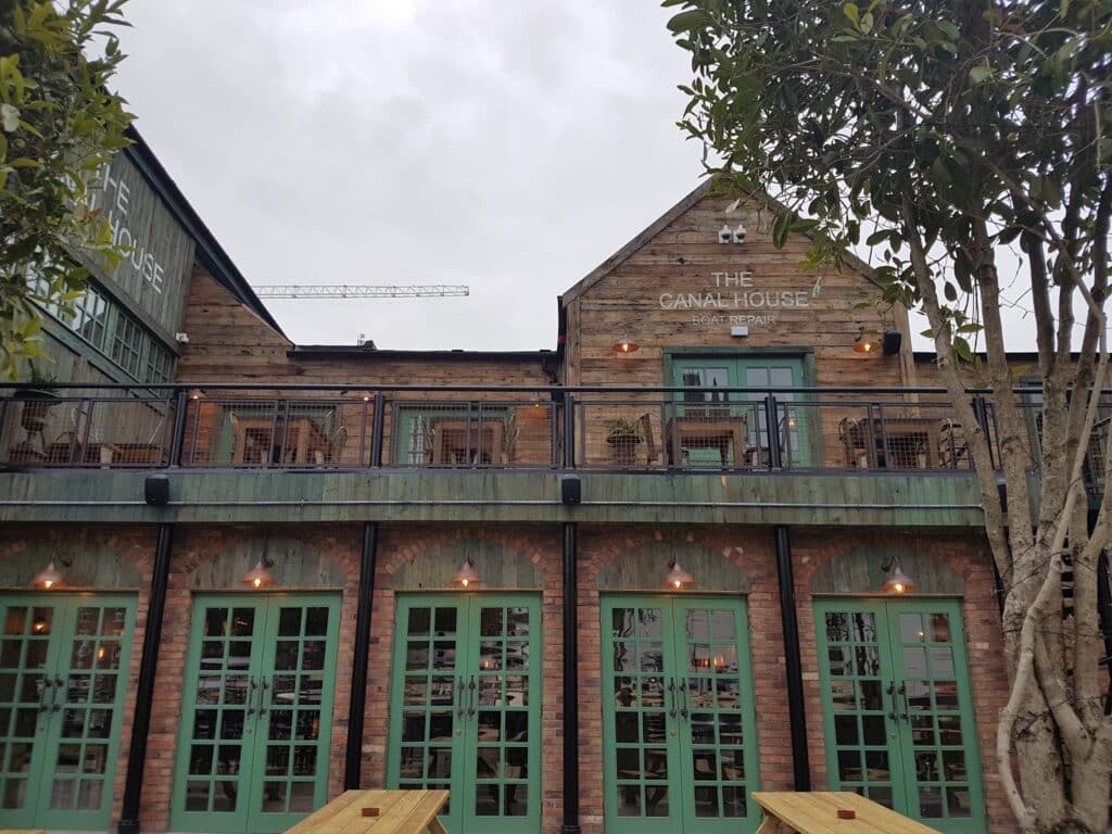 A two-storey rustic building houses The Canal House pub. The lower level has green-framed glass doors, while the upper level features wooden walls, a balcony with tables and chairs, and a sign reading "THE CANAL HOUSE." A tree partially frames the right side.