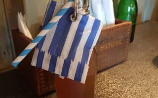 A glass bottle with a flip-top lid, filled with a light brown drink, is topped with a blue and white striped paper straw and matching serviette. The bottle is on a textured black surface next to cutlery, a menu, and a green glass bottle in the background.