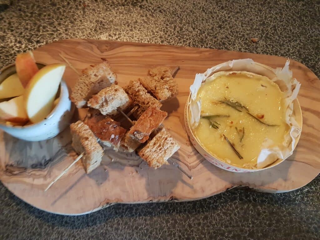 A wooden serving board with baked Camembert cheese garnished with rosemary, cubes of brown bread on skewers, and a small bowl of apple slices, set on a textured grey surface.