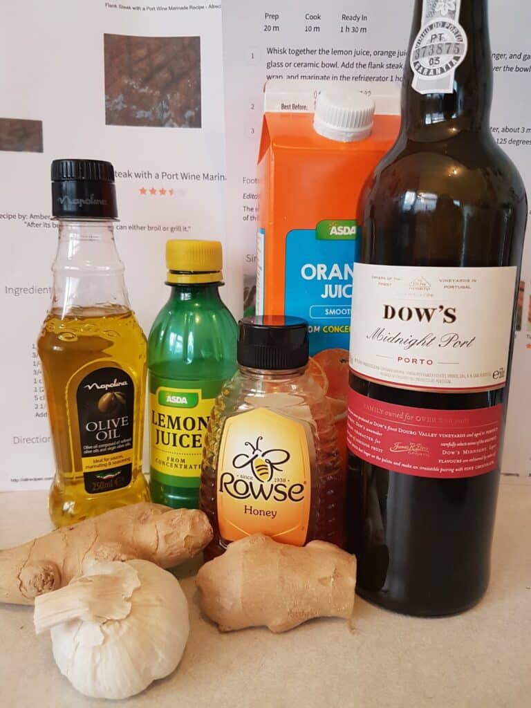 A group of cooking ingredients on a worktop: a bottle of olive oil, lemon juice, orange juice carton, bottle of port wine, jar of honey, and pieces of fresh ginger. A recipe with text is partially visible in the background.
