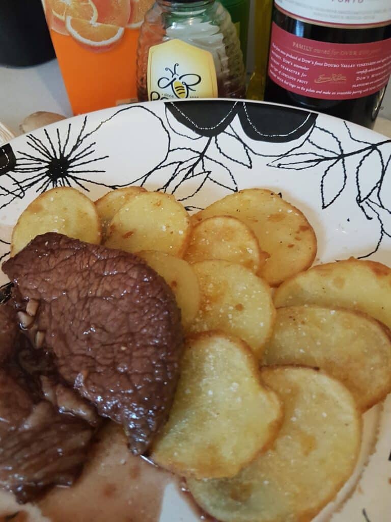 A plate with two slices of cooked beef in dark sauce next to neatly arranged, round golden potato slices. In the background are a bottle of honey, a bottle of olive oil, a wine bottle, and an orange mug. The plate has a black floral pattern.
