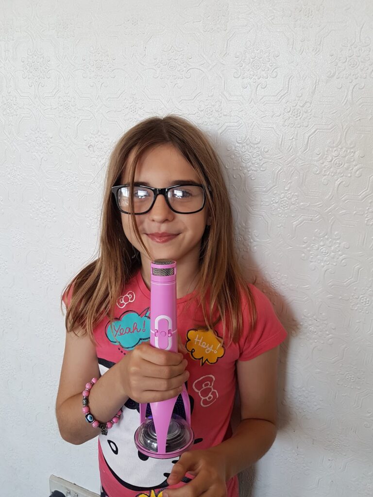 A young girl with straight brown hair and glasses stands against a textured white wall, smiling. She wears a pink shirt with colourful patches, a beaded bracelet, and holds a pink toy microphone with both hands.