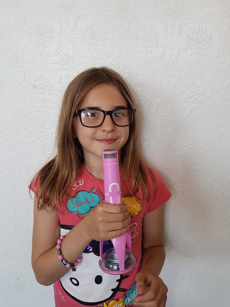 A girl with long light brown hair and glasses stands against a textured white wall, smiling slightly. She wears a pink Hello Kitty shirt with colourful designs and holds a pink toy microphone with both hands. She also wears a beaded bracelet.