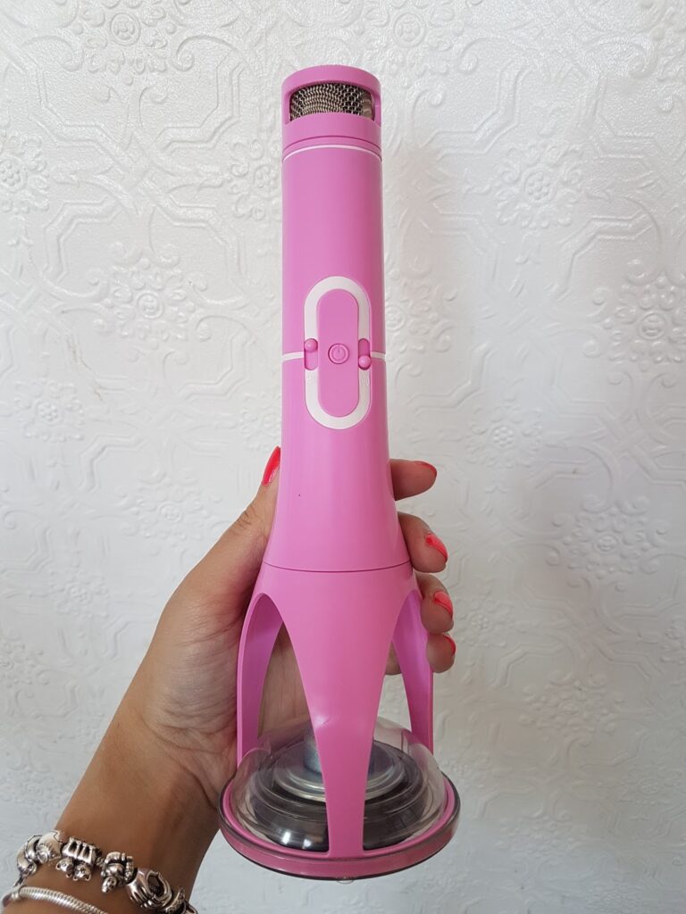 A hand with pink painted nails and a silver bracelet holds a pink handheld microphone with a built-in stand. The background features a white textured floral-patterned wall.