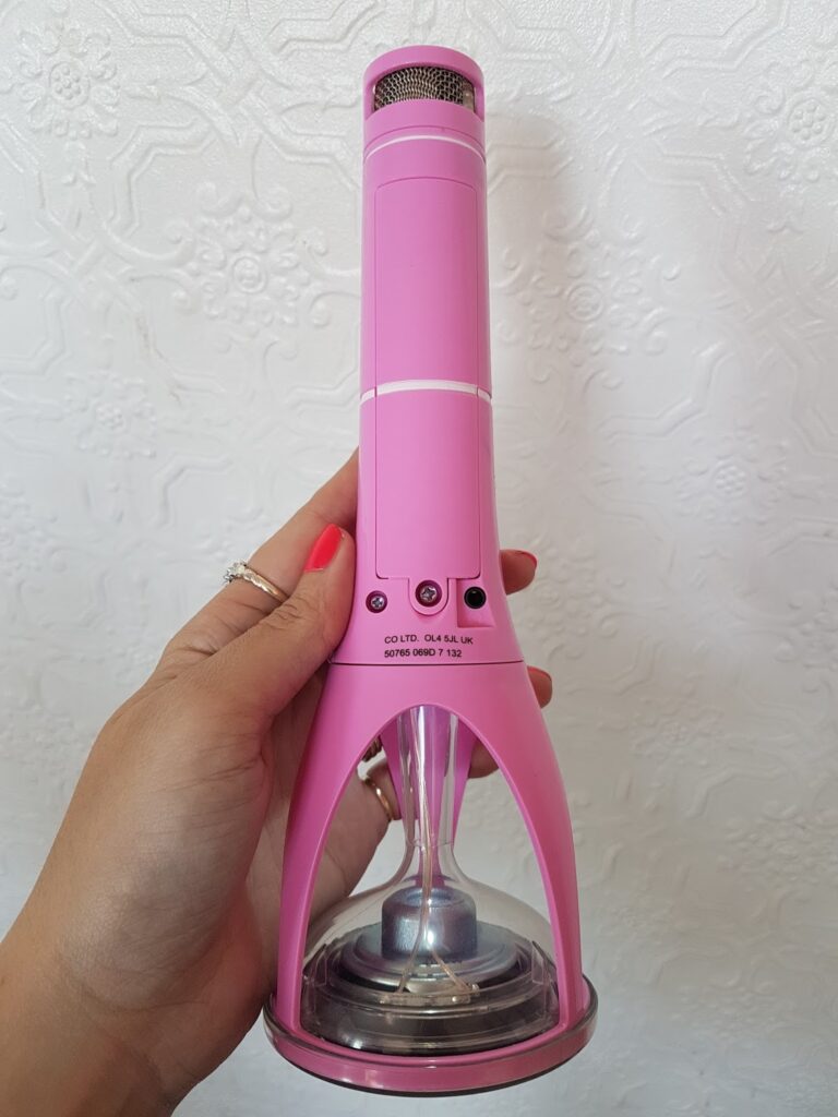 A hand with red nail varnish holds a bright pink handheld microphone, which is attached to an unusual transparent base. The background is a white textured wall with a subtle floral pattern. The device has visible screws and printed product information.