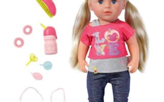 A blonde doll with blue eyes wears a pink shirt with “LOVE” and cartoon graphics, blue jeans, and pink shoes. Accessories beside her include a pink hairbrush, a bottle, two teething rings, a dummy, two bangles, and a pink oval comb.