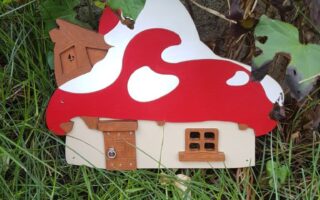 A small, whimsical house with a red and white mushroom-shaped roof sits among tall green grass and leafy plants. The house has a tiny wooden door and windows, resembling a fairy or gnome home nestled in a garden.
