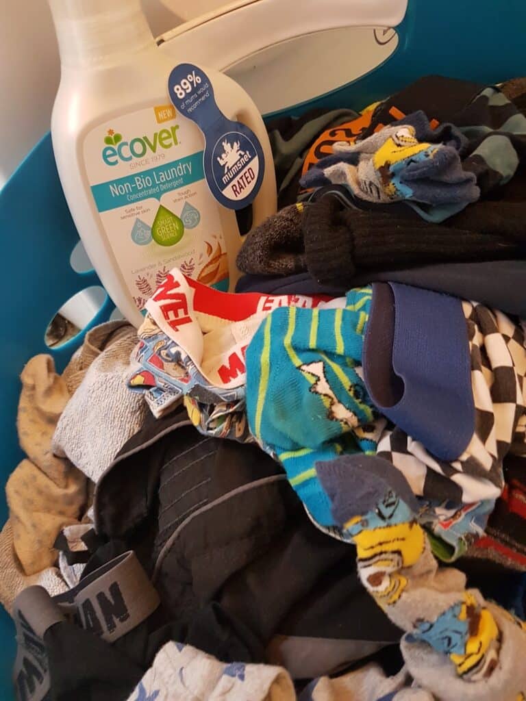 A blue laundry basket filled with mixed clean clothes, including colourful socks and underwear, with a white bottle of Ecover Non-Bio Laundry detergent placed on top. The detergent label shows “88% less plastic” and “Mother & Baby Rated”.