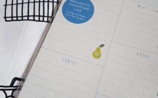 A magnetic shopping list pad divided into sections—fruit & vegetables, larder, meat & seafood, dairy, and home—sits on a white surface with a black line drawing of a shopping trolley. The list has simple illustrations, like a pear and a teapot.