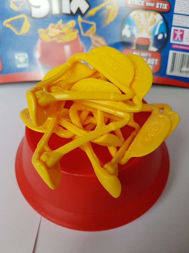 A stack of yellow plastic sticks, shaped with paddle-like ends, balances on a red bowl. The background features a blue game box with "Stix" in bold white letters and game instructions, partially visible.