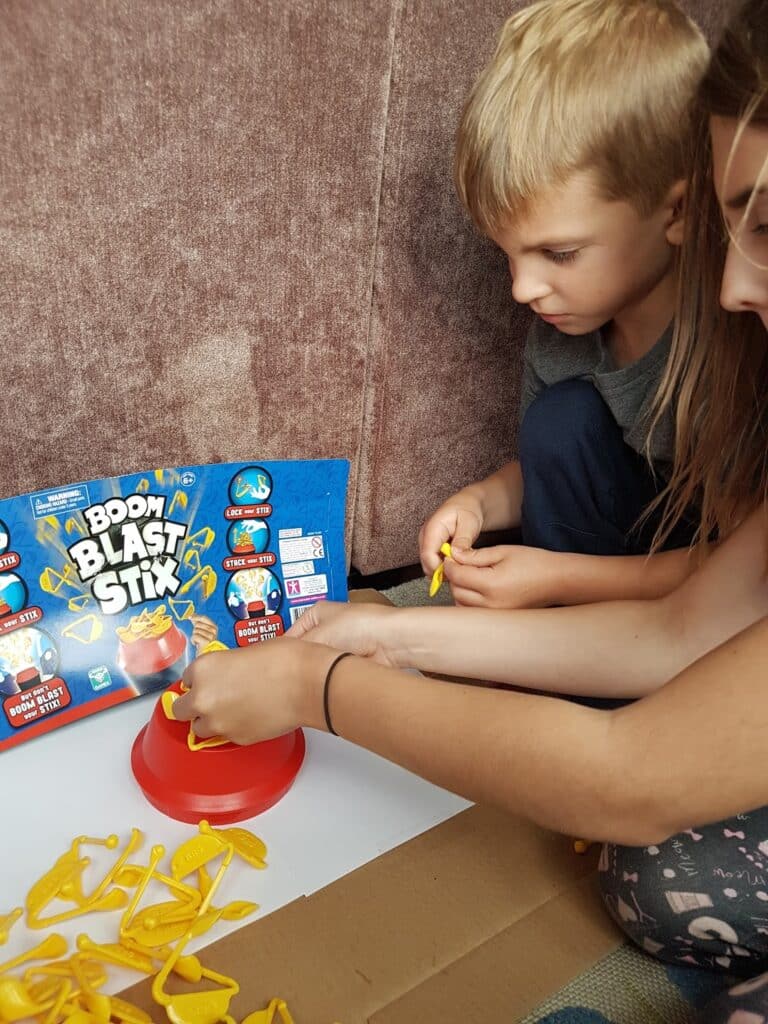 Two children sit on the floor playing the game Boom Blast Stix. The boy watches intently whilst the girl places a yellow plastic stick onto a circular red base. Many yellow sticks are scattered around them next to the game box.