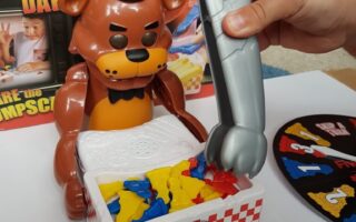 A hand uses a grey plastic claw to pick up coloured pieces from a box in front of a brown bear toy with a black hat and bow tie. The game is themed after Five Nights at Freddy’s, as shown by the box art and logo in the background.