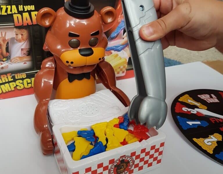 A hand uses a grey plastic claw to pick up coloured pieces from a box in front of a brown bear toy with a black hat and bow tie. The game is themed after Five Nights at Freddy’s, as shown by the box art and logo in the background.